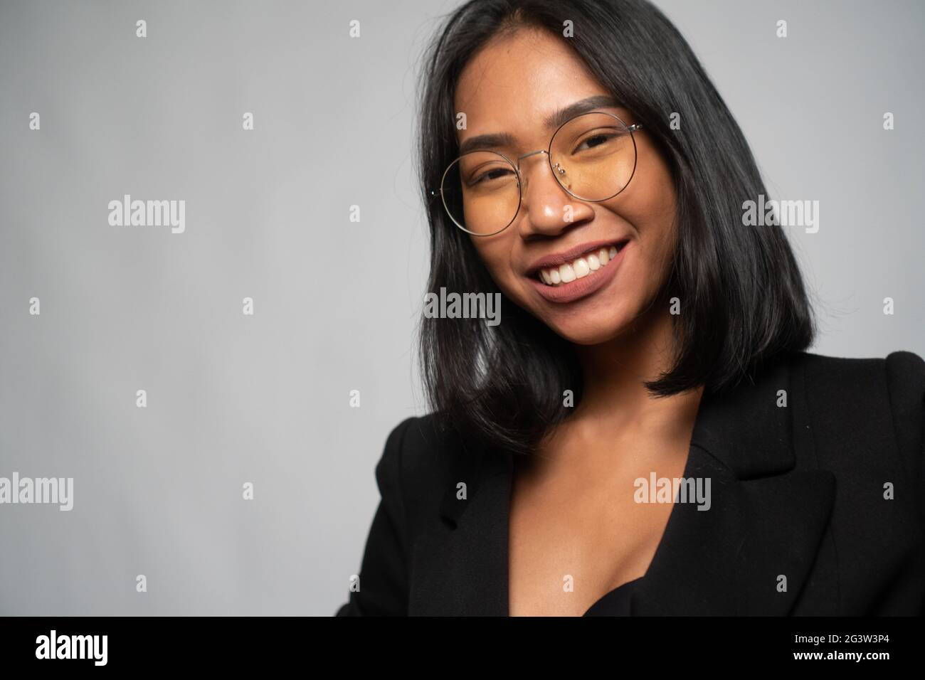 Attraente giovane donna asiatica in elegante blazer nero e vetri isolati su parete grigio chiaro Foto Stock