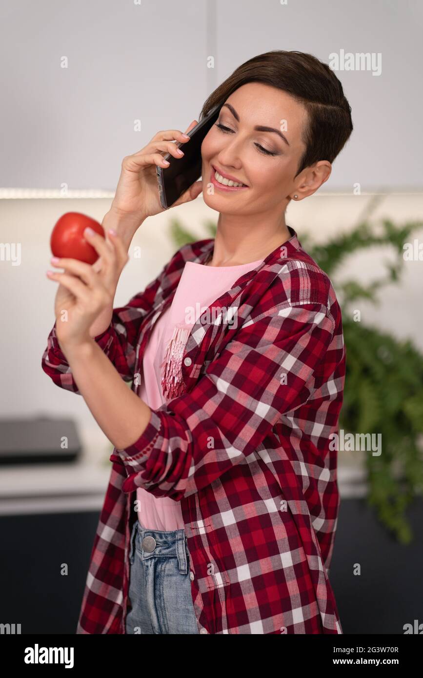 Scelta del pomodoro fresco mentre parla sulla casalinga del telefono con una pettinatura corta. Pranzo di cucina per la famiglia nel moderno angolo cottura Foto Stock