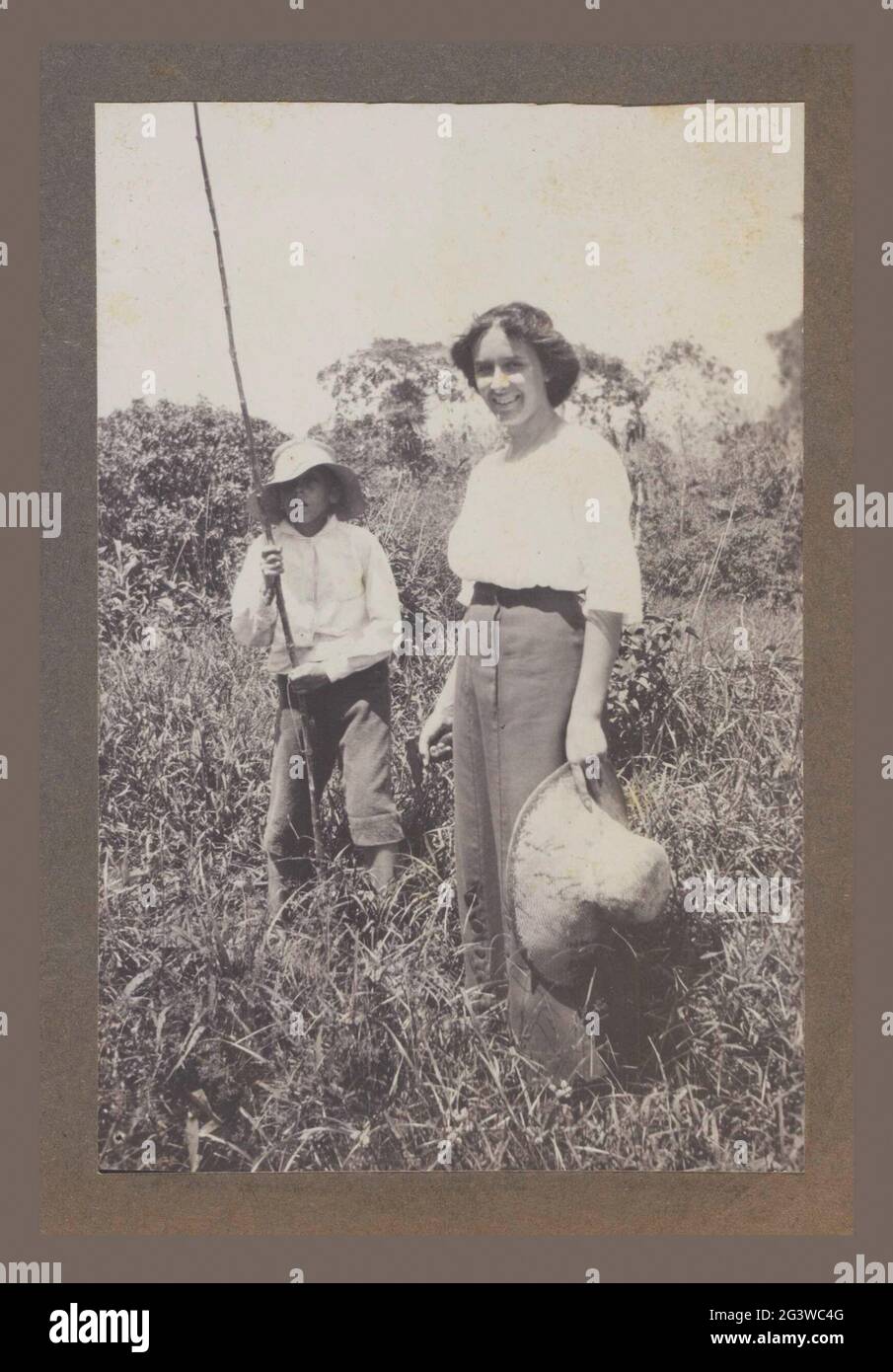 Maria Gonggrijp, in piedi con il cappello in mano. Maria gonggrijp, sorridente e in piedi in erba con il cappello da sole in mano. Dietro di lei un ragazzo con un bastone (asta?). Probabilmente su Plantage Clevia. Parte dell'album fotografico sulla vita della famiglia Gonggrijp in Suriname intorno al 1912. Foto Stock