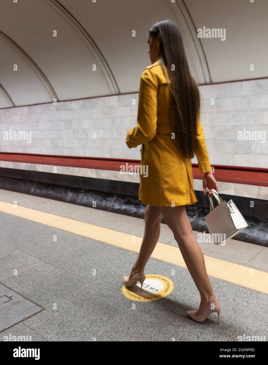 Vista posteriore di una passeggiata sulla piattaforma della metropolitana bella ragazza giovane con lunghe e belle gambe con un cappotto di primavera giallo e un bianco Foto Stock