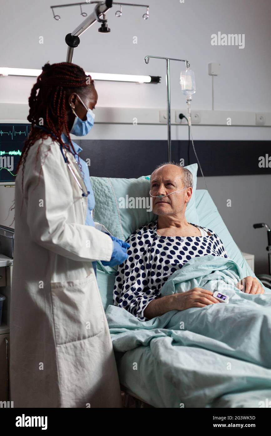 Medico afroamericano che indossa maschera chirurgica, in sala d'ospedale durante la consultazione di malato malato uomo anziano, respirando con l'aiuto di tubo di prova e fleboclisi iv attaccato. Foto Stock