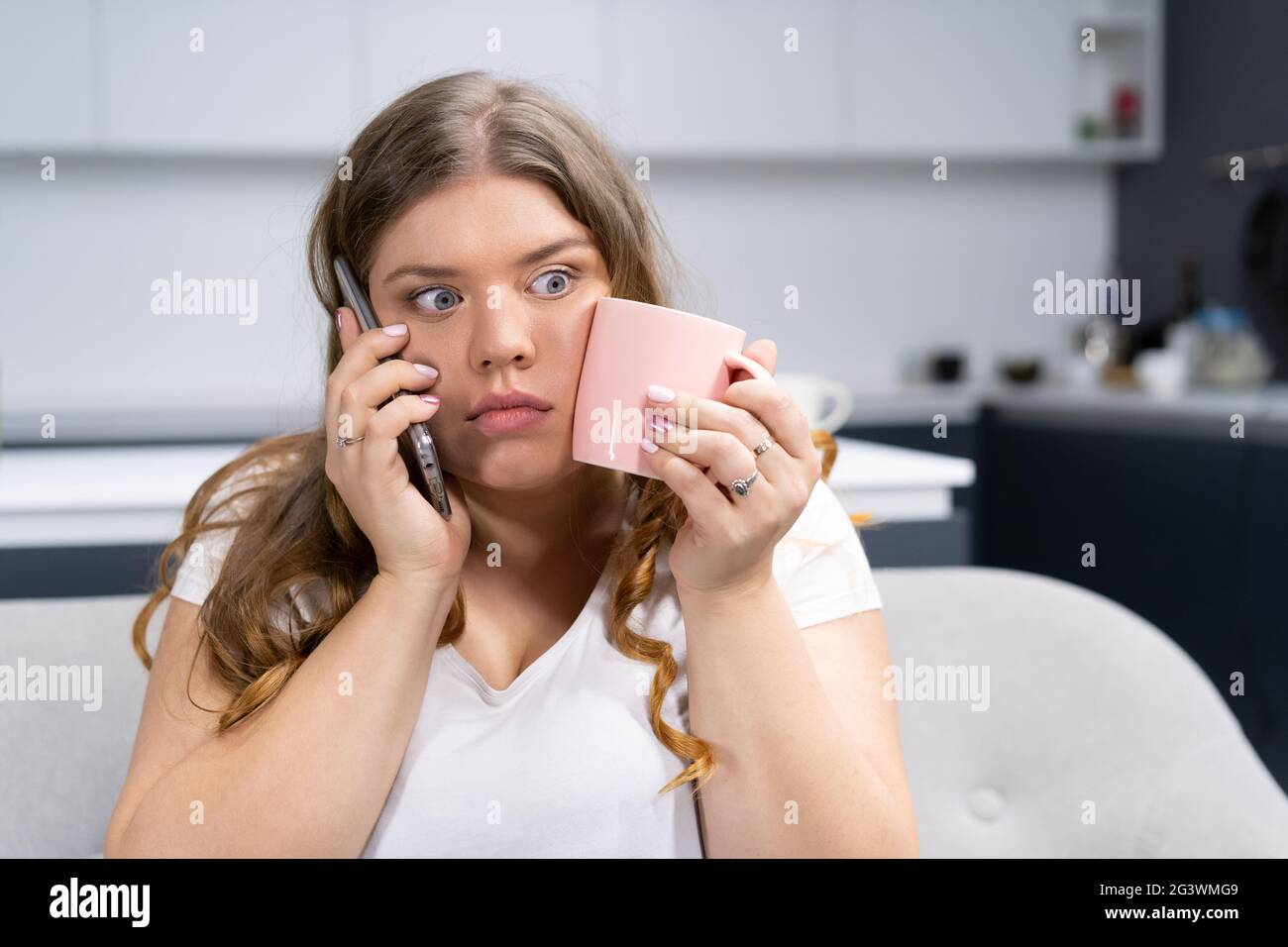 Scioccato o sorpreso guardare sovrappeso giovane donna che parla al telefono con il capo o gli amici tenendo una tazza con acqua fredda touchin Foto Stock