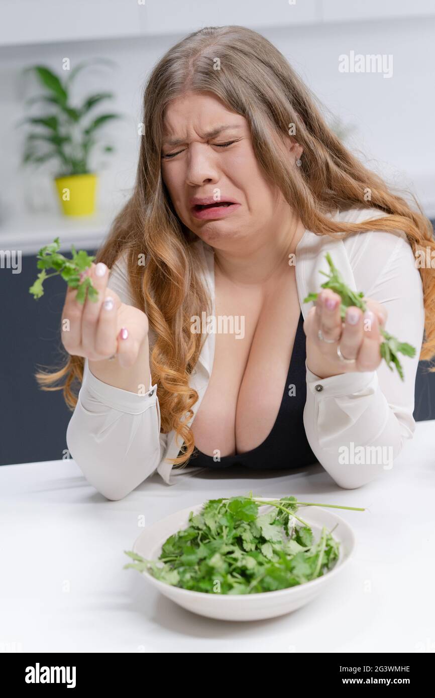 Fat ragazza non felice tenendo fresco insalata casting. Giovane donna dal corpo curvo con lunghi capelli biondi seduta sulla cucina moderna. Dieta A. Foto Stock