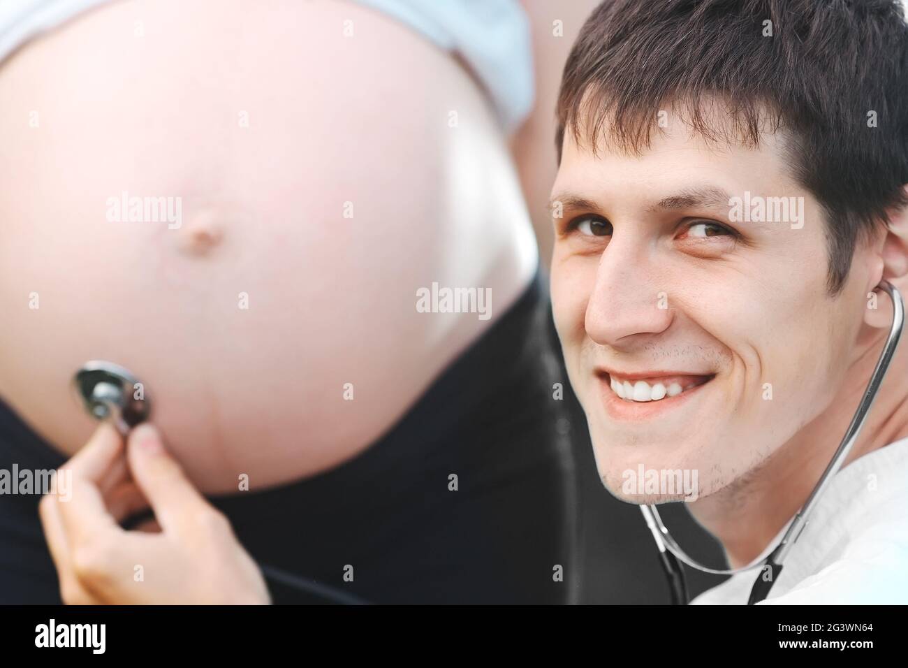 Il giovane medico ascolta lo stomaco della donna incinta e sorride alla telecamera. Per avere una gravidanza sana. Foto Stock