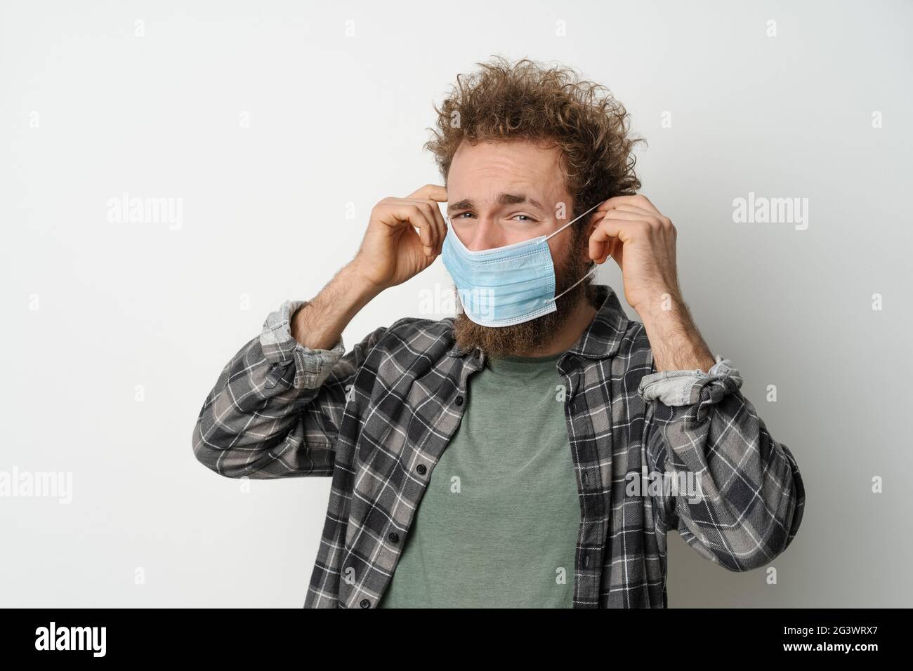 Disgustedly sotto la tregua mette sulla sua faccia una maschera medica protettiva per proteggere il coronavirus, con capelli ricci, giovane uomo che indossa Foto Stock