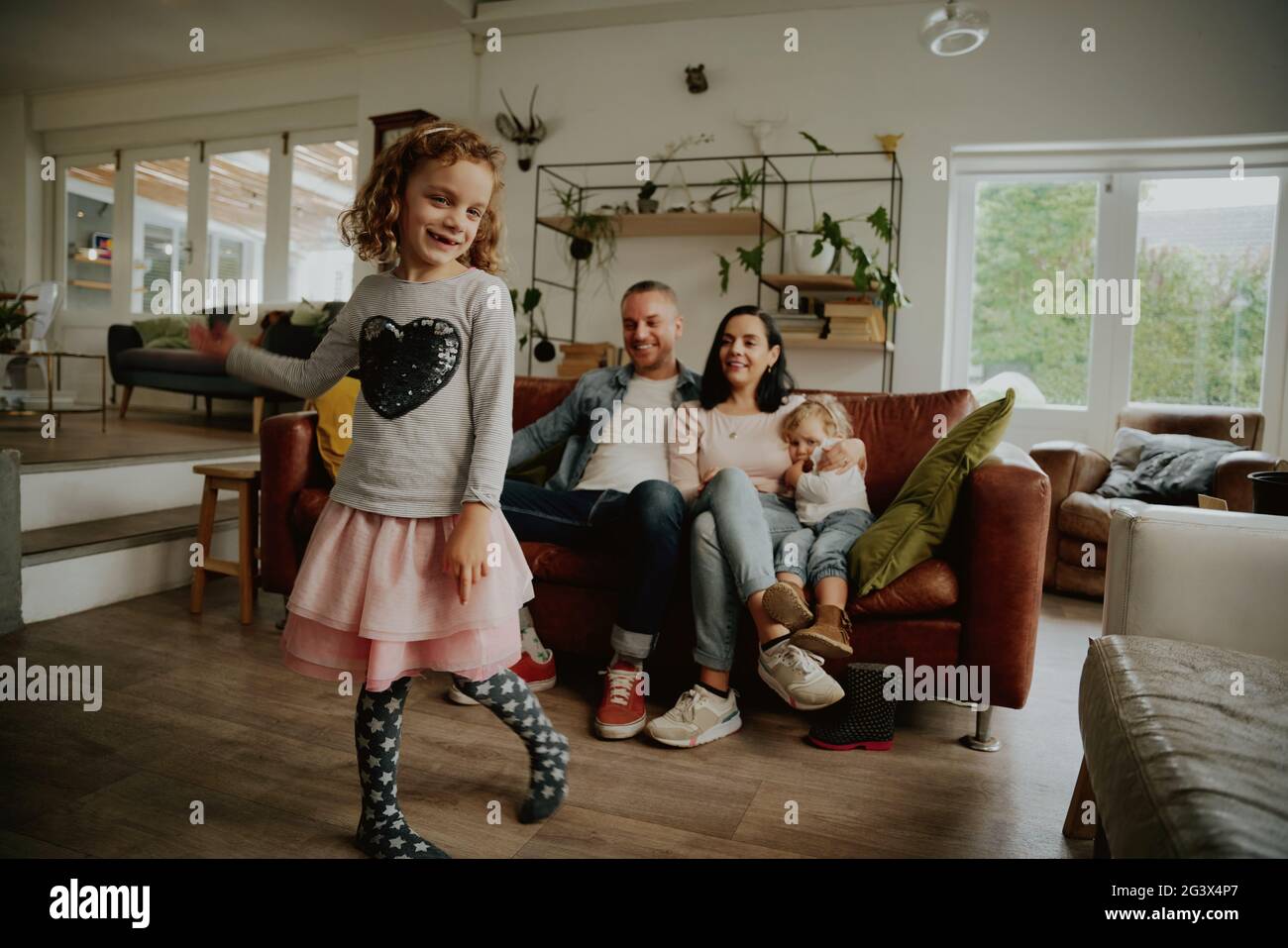 Ragazza carina che danzano con i genitori e la sorella più giovane che guarda seduto sul divano a casa Foto Stock