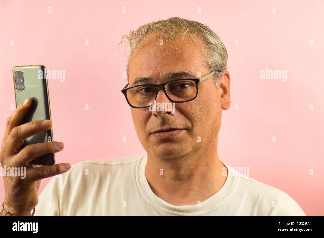primo piano di un uomo dai capelli grigi con occhiali che tengono una mobil guardando dritto su uno sfondo rosa Foto Stock