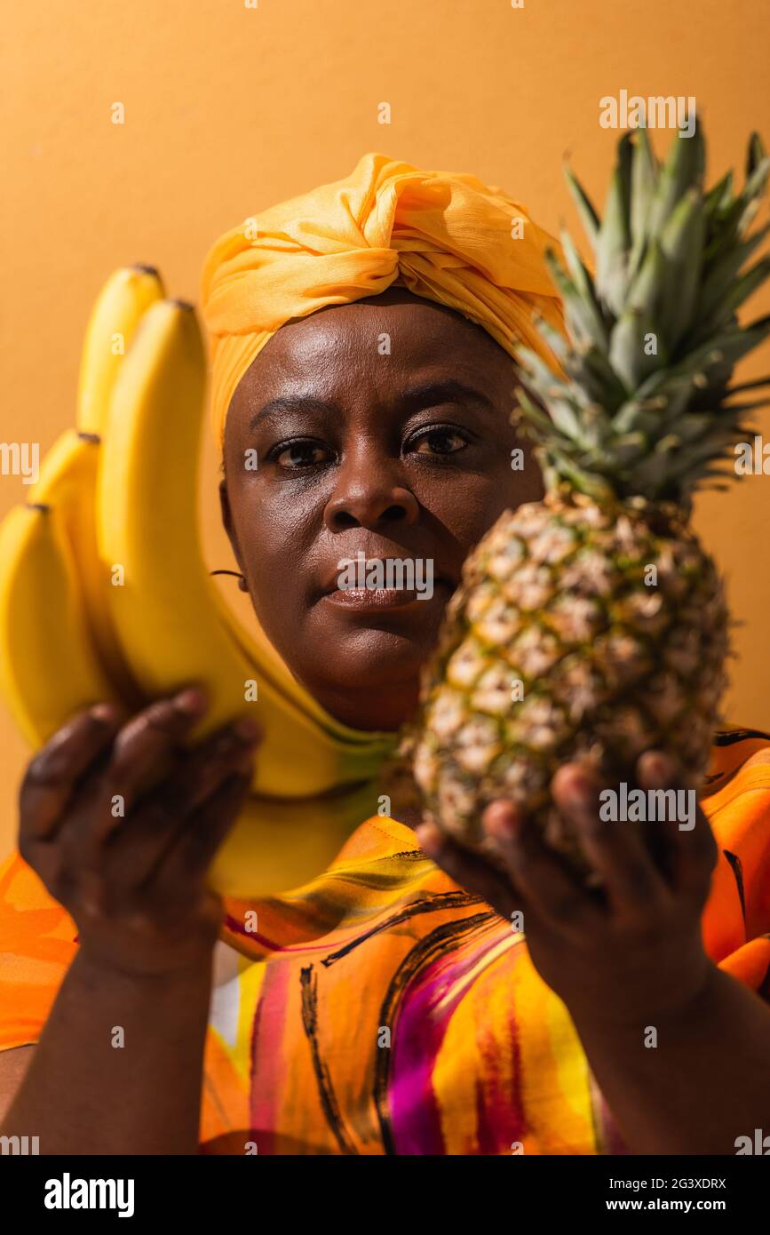 donna afroamericana in turbano giallo e vestito colorato tenendo ananas e banane su arancio Foto Stock