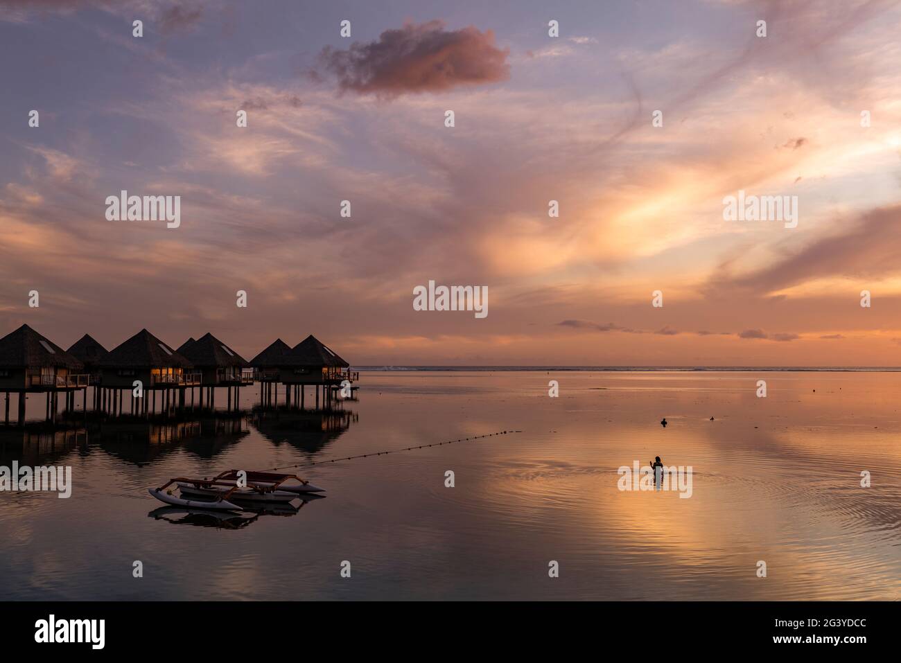 Silhouette di donna in acqua accanto alla canoa Outrigger e bungalow sull'acqua del Tahiti Ia ora Beach Resort (gestito da Sofitel) al tramonto, Foto Stock