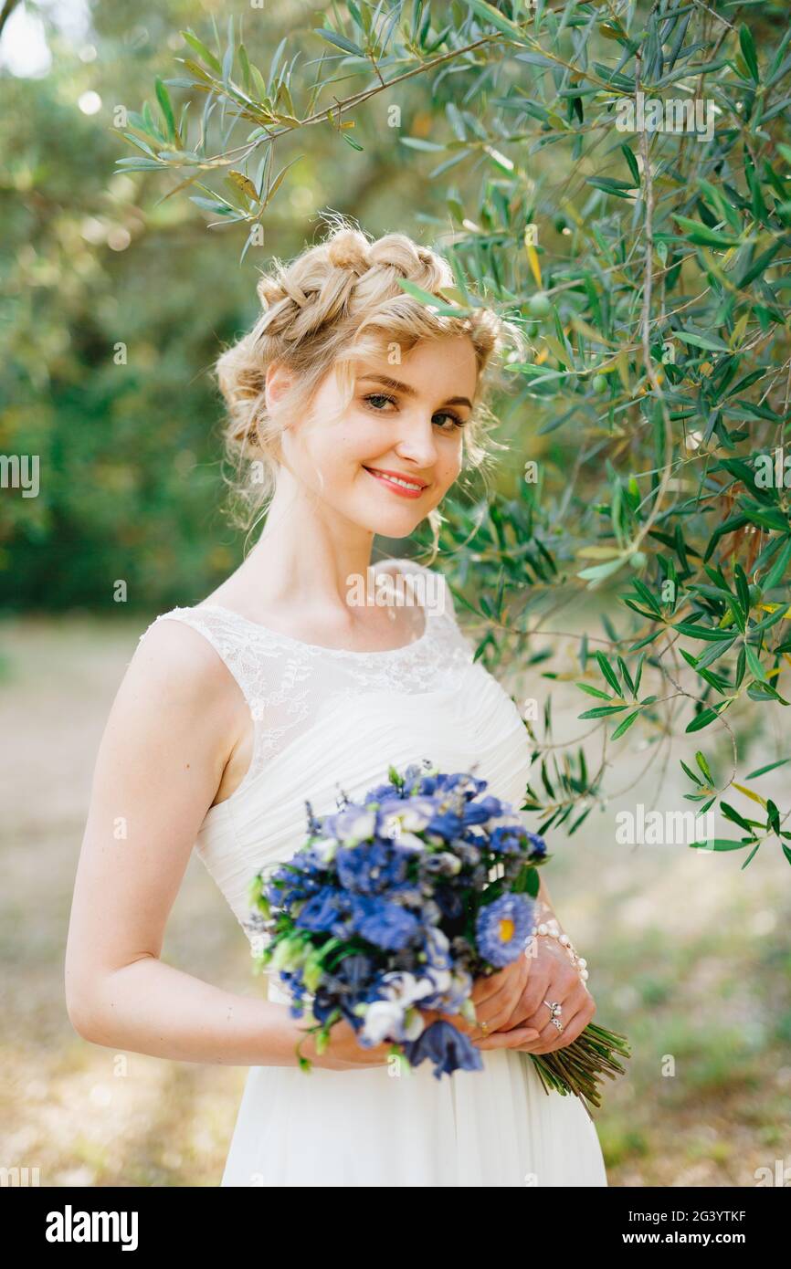 Una sposa tenera con un bouquet di fiori blu in le sue mani sono in piedi da rami di olive verdi in un boschetto e sorrisi Foto Stock