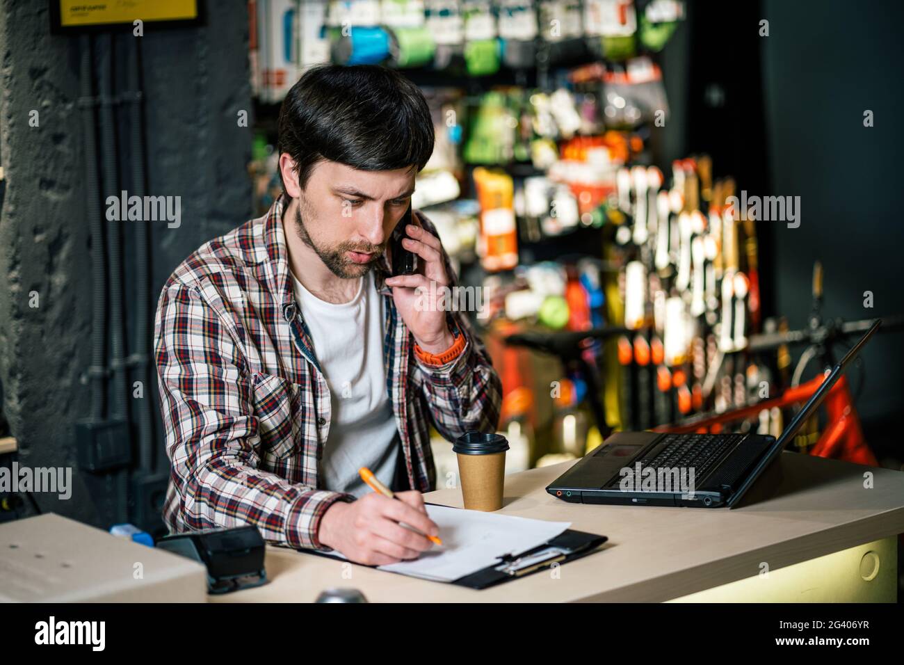 Proprietario del negozio di biciclette al lavoro. Il dipendente del negozio effettua un ordine tramite telefono cellulare al tavolo vicino al laptop nel negozio di articoli da viaggio e sportivi. Foto Stock