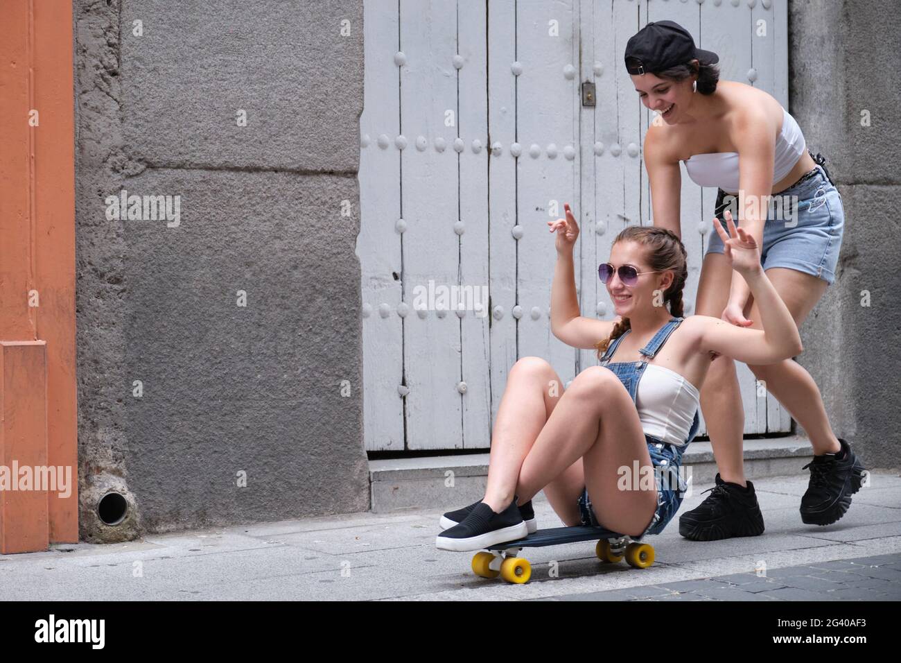 Una donna che spinge la sua amica da dietro su uno skateboard. Due amiche che si divertono in strada. Foto Stock