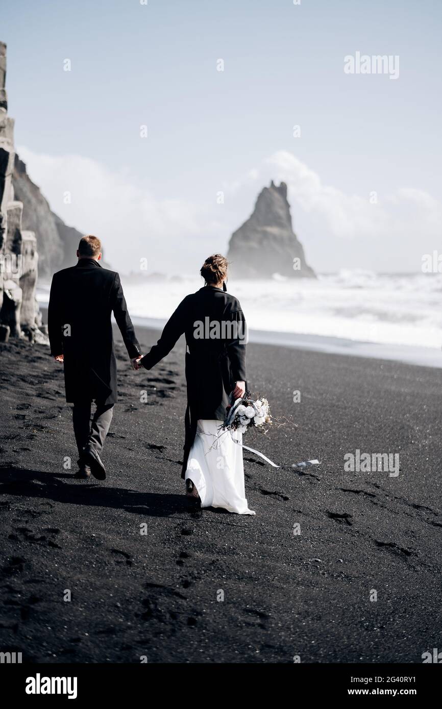 Destinazione Islanda matrimonio. Una coppia di nozze cammina lungo la spiaggia di sabbia nera di Vik, vicino alla roccia di basalto, sotto forma di PIL Foto Stock