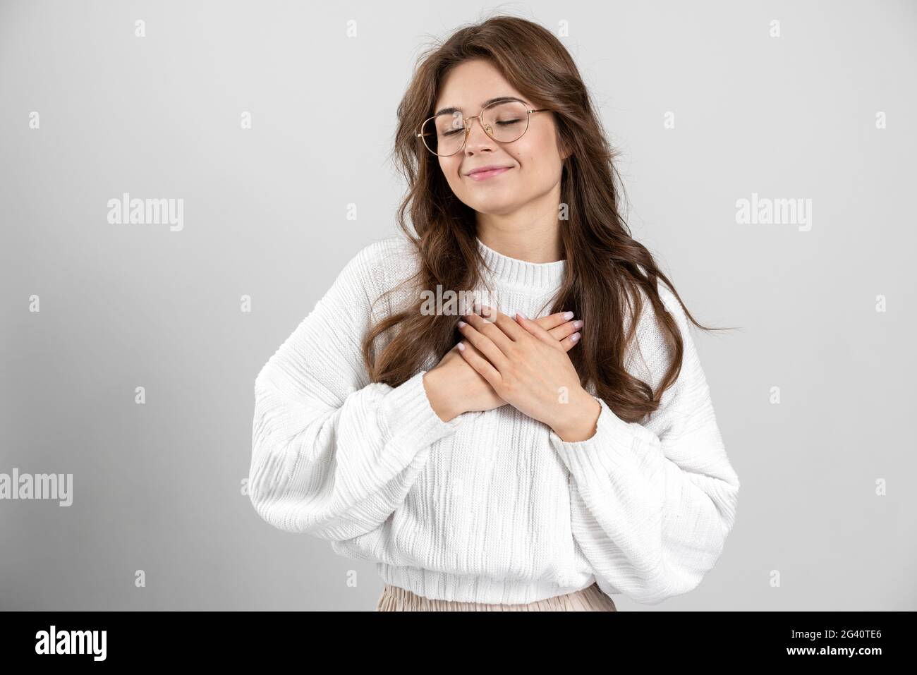 Ritratto di una ragazza carina e tenera, con gli occhiali, ha messo le mani al suo cuore, mostra un segno dal cuore. Bella ragazza gesturing su uno sfondo grigio Foto Stock