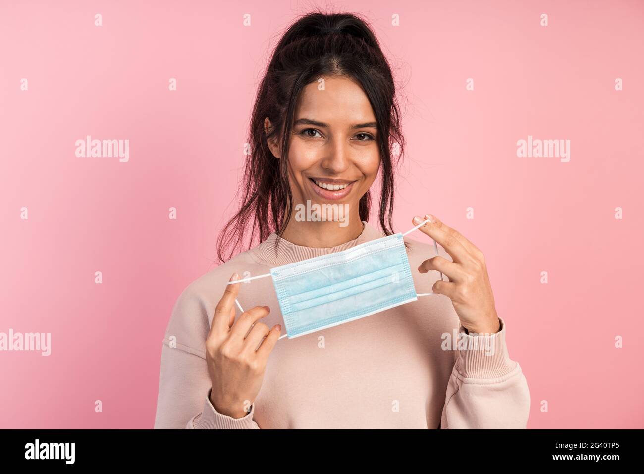 Cute, ragazza attraente tiene una maschera protettiva nelle sue mani e la mette sopra. Bella bruna sorridente isolata su sfondo rosa. Foto Stock