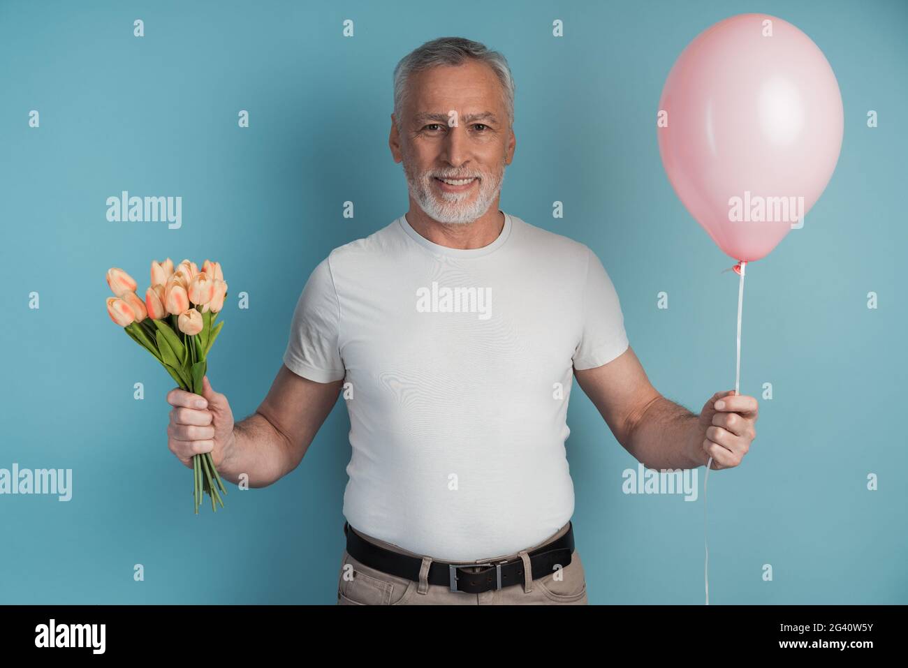 Gallante, l'uomo anziano tiene i fiori e palloncino rosa in mano. Uomo sorridente con capelli grigi su sfondo blu, posto per il testo, copia spazio. Foto Stock