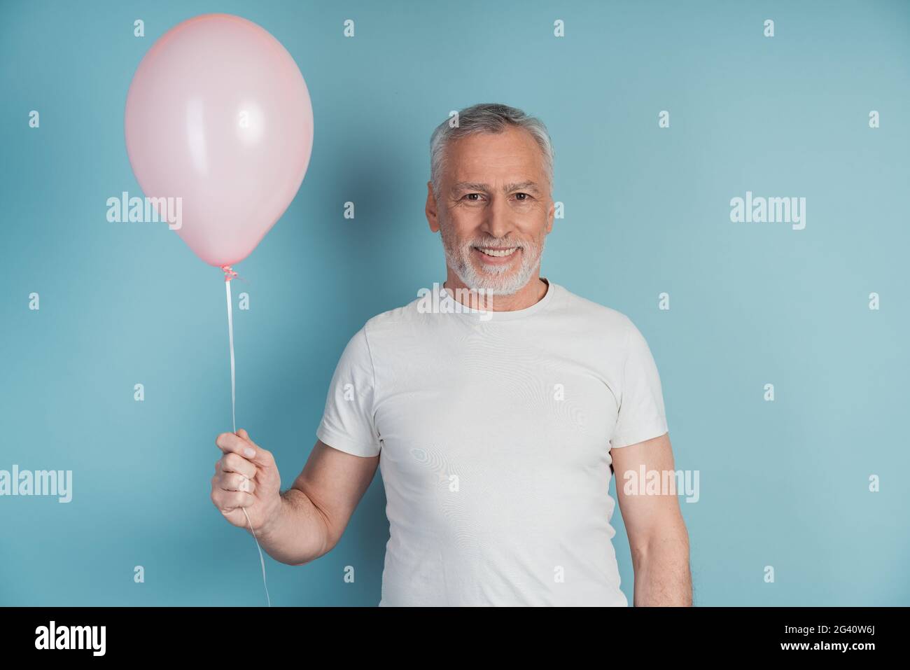 Felice uomo in pensione in t-shirt bianca che tiene il palloncino rosa mentre si posa in studio. Uomo anziano, sorridente su sfondo blu, copia spazio, posto per il testo Foto Stock