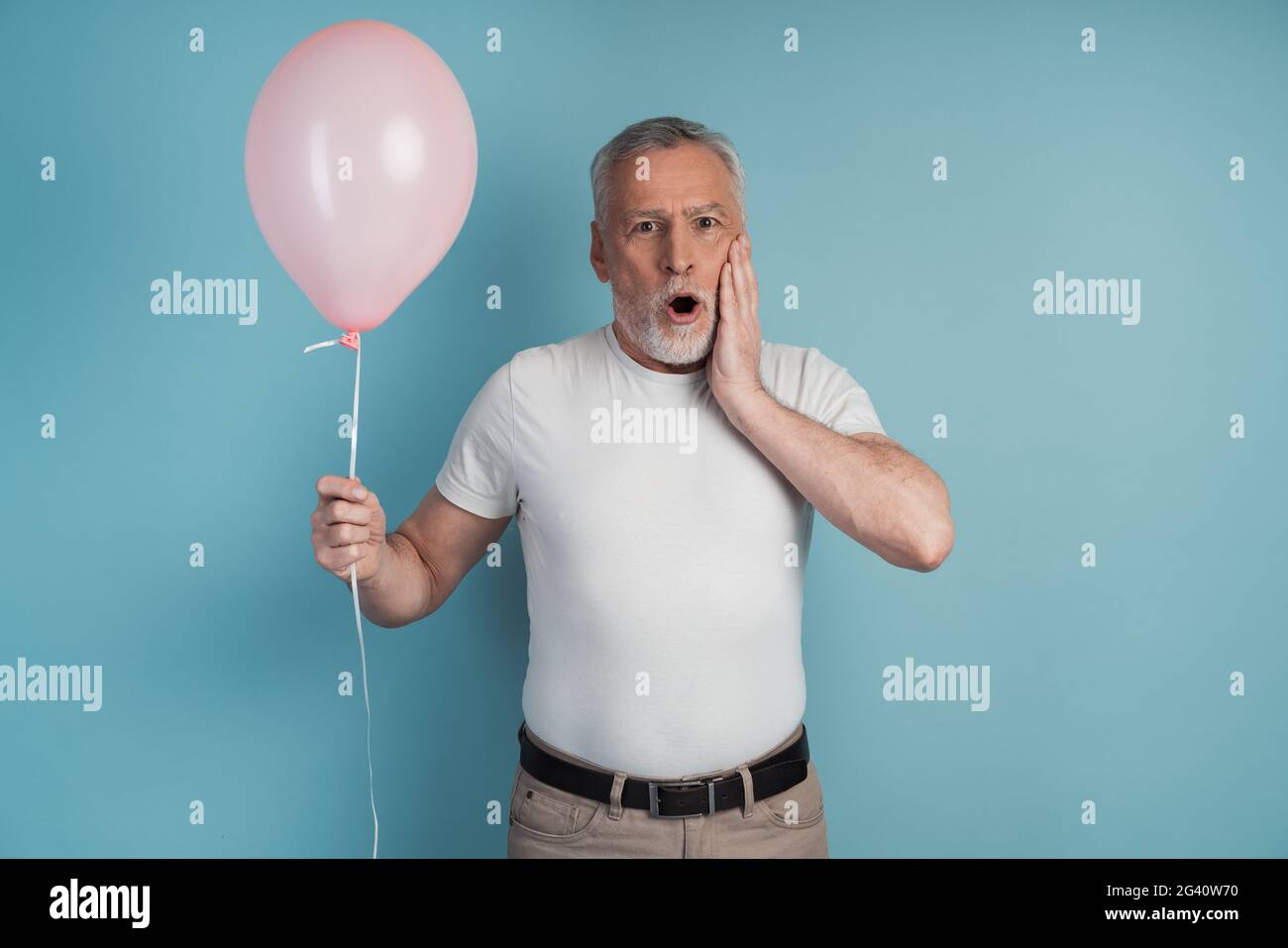 Uomo anziano sorpreso che tiene una palla rosa in mano. Uomo con capelli grigi su sfondo blu, posto per il testo, copia spazio. Foto Stock