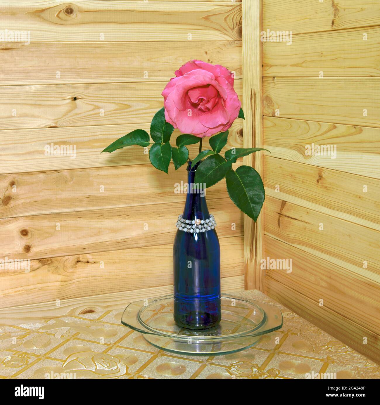 Una rosa nella bottiglia blu sul tavolo da cucina nell'angolo interno sullo sfondo della parete di rivestimento in legno. Foto Stock