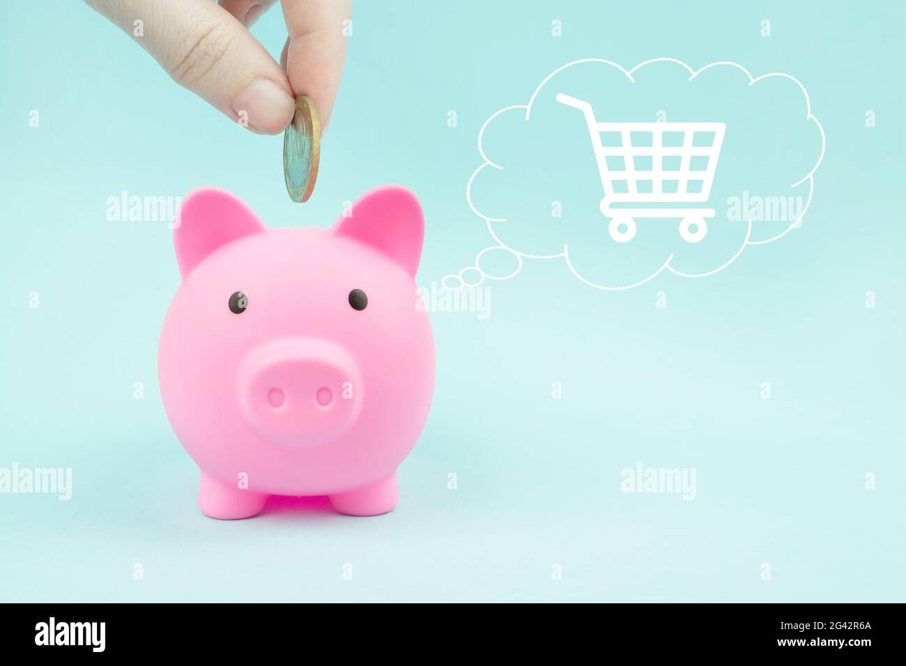 Mano che mette una moneta in una banca piggy con carrello di shopping ologramma digitale in nube pensato sopra la sua testa su sfondo blu. Risparmio di denaro, finanziario p Foto Stock