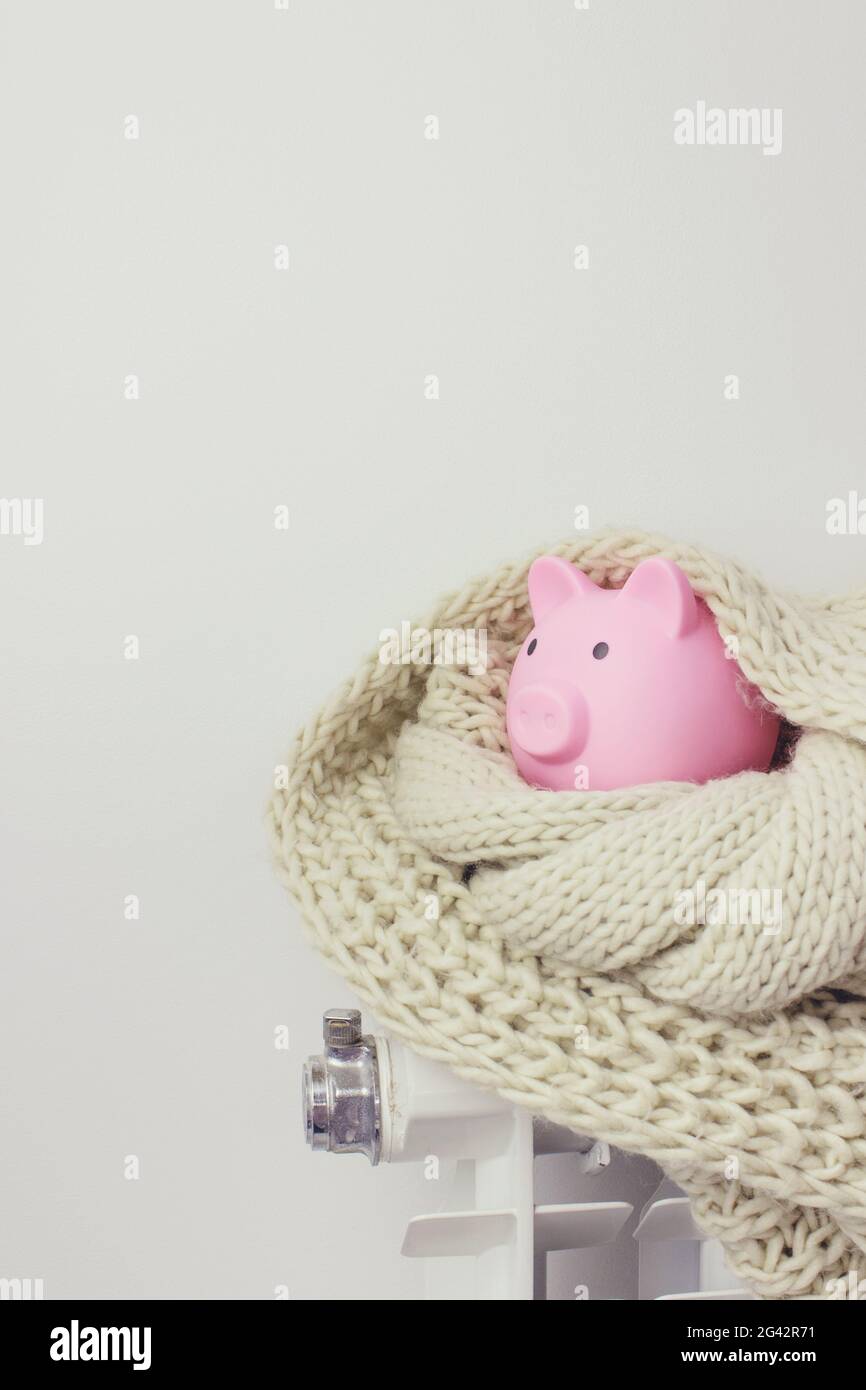 Risparmio denaro ricchezza e concetto finanziario. Banco di piggy e sciarpa sul radiatore di riscaldamento su sfondo grigio. Concetto di stagione di riscaldamento. Foto Stock