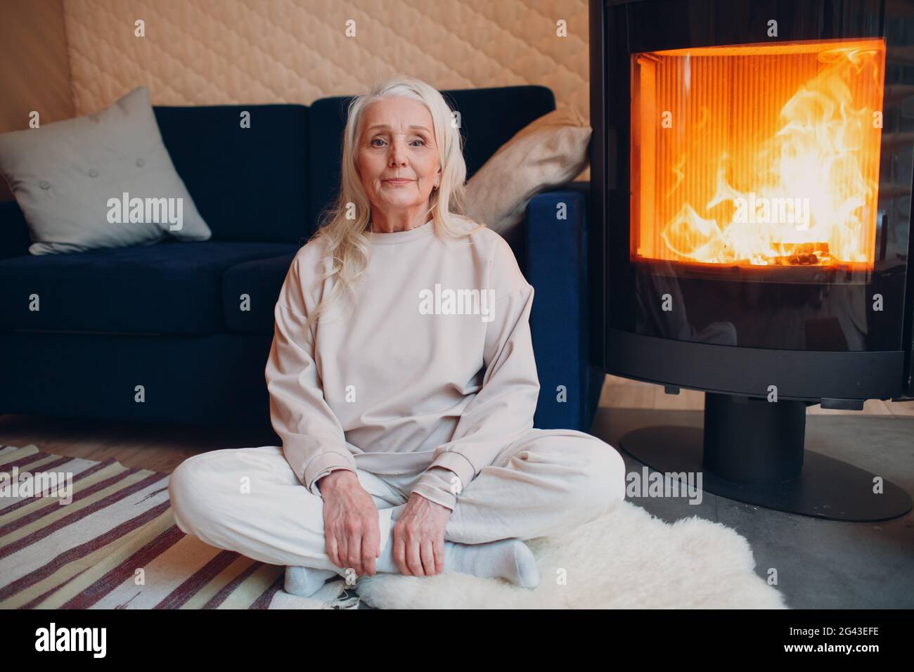 Donna con capelli grigi mature seduta su divano in soggiorno con camino Foto Stock