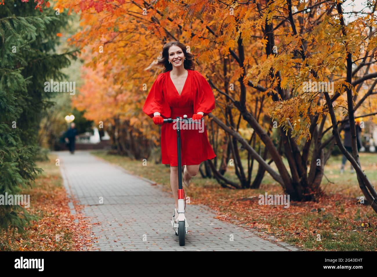 Giovane donna con scooter elettrico in abito rosso al parco cittadino d'autunno Foto Stock