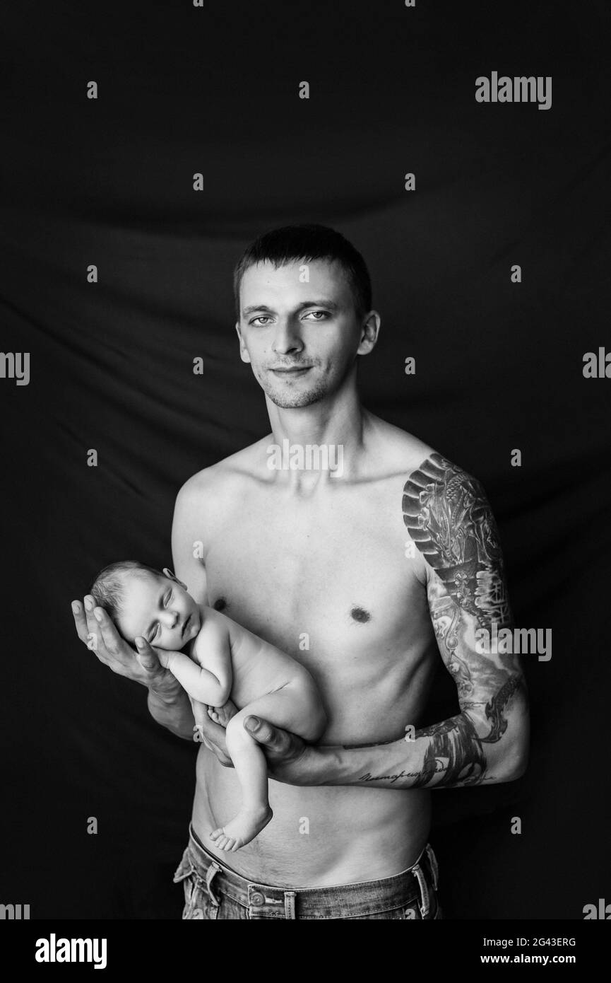 Il padre con il tatuaggio sul braccio tiene il suo neonato su uno sfondo bianco e nero Foto Stock