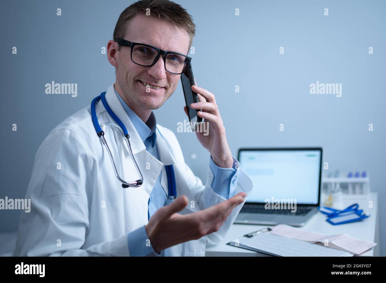 Medico europeo in cappotto medico bianco e bicchieri consulta il paziente sul telefono cellulare in ufficio clinica, seduto al tavolo con il computer portatile. Foto Stock