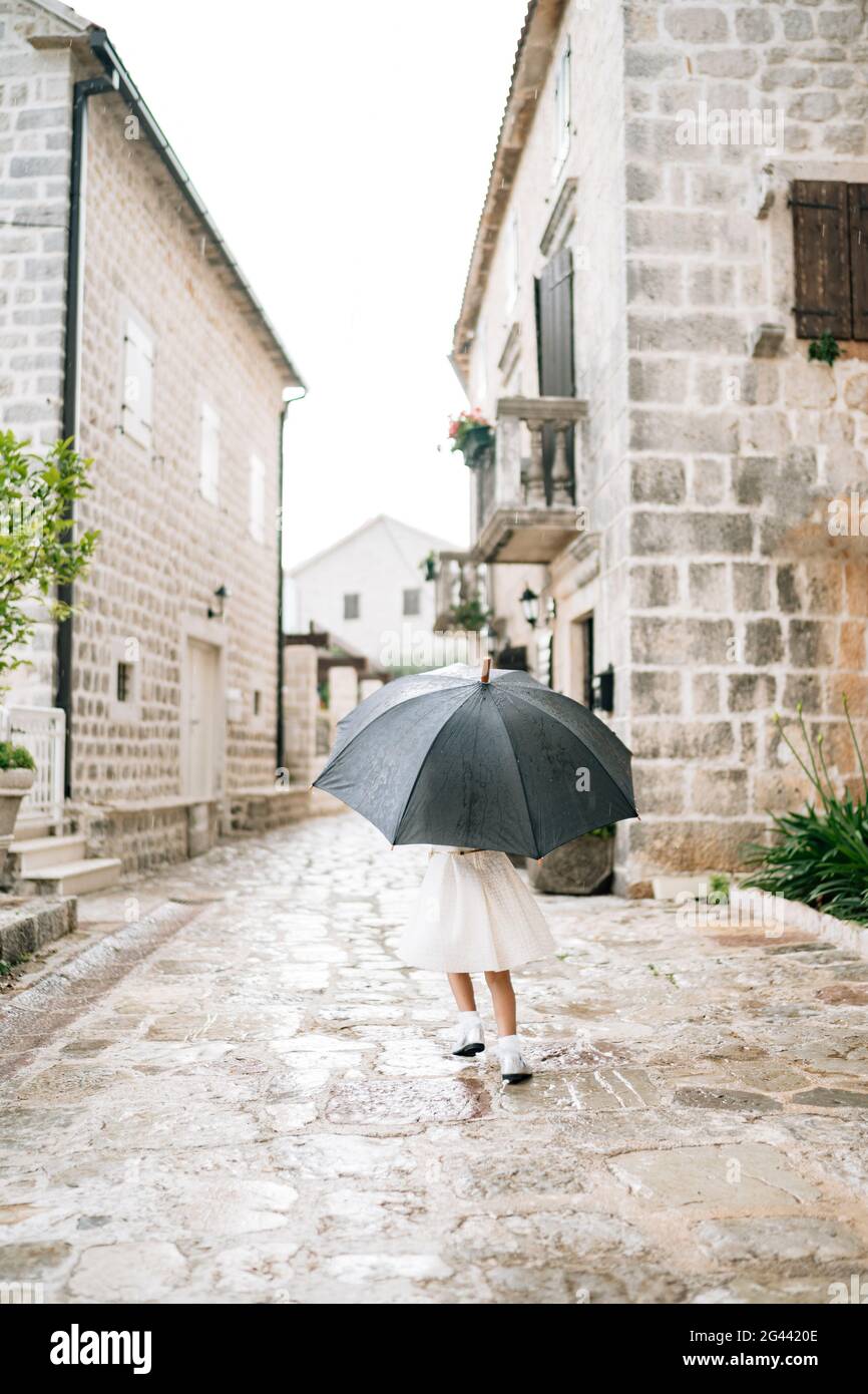 Il bambino si posa con un ombrello. Una bambina in un abito si trova fuori sotto un ombrello nero durante la pioggia. Foto Stock