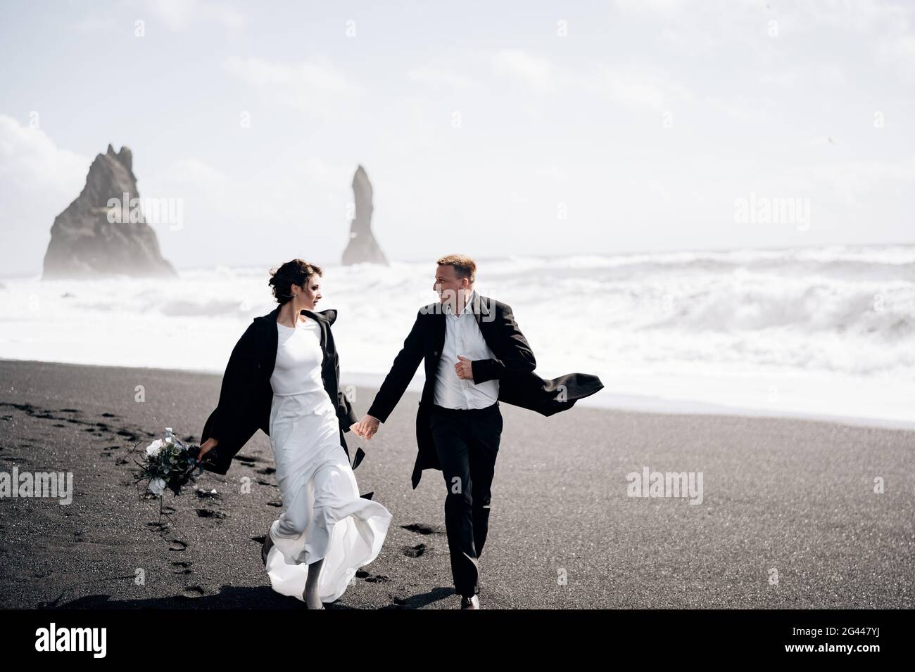 Destinazione Islanda matrimonio. La coppia di nozze corre lungo la spiaggia sabbiosa nera di Vik, vicino alla roccia basaltica, sotto forma di pi Foto Stock
