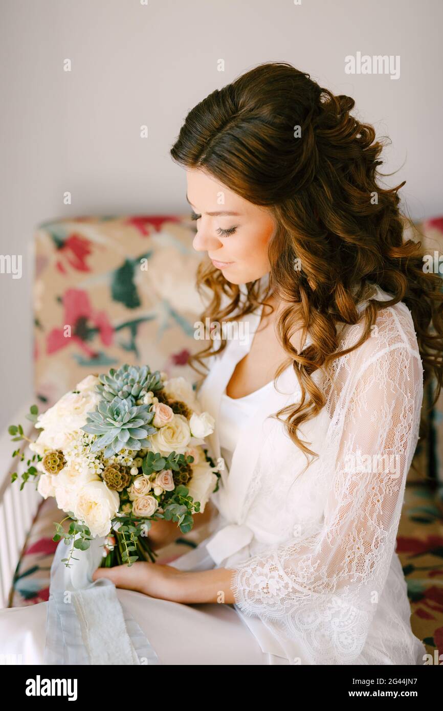 Una sposa in un abito di pizzo si siede su un divano in una camera d'albergo e tiene un bouquet di nozze nelle sue mani, da vicino Foto Stock