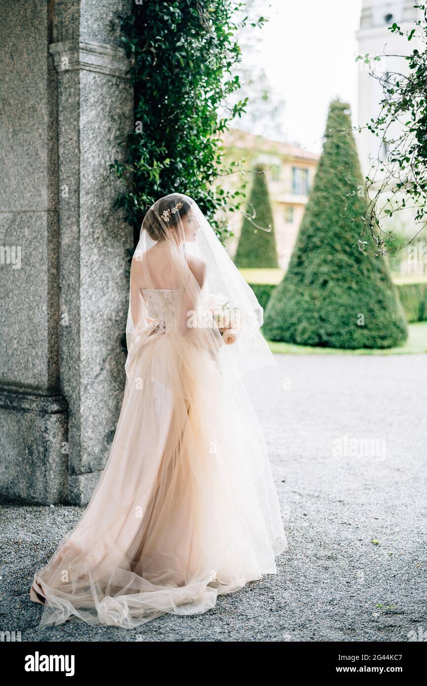 Sposa in un abito da sposa con un velo e un bouquet di fiori si trova  vicino all'arco di un vecchio edificio di fronte a un giardino verde. L  Foto stock -