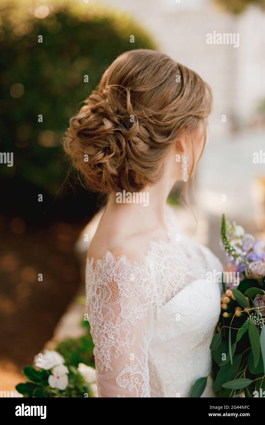 Una sposa con una bella acconciatura in un abito in pizzo si erge con un bouquet e guarda a lato, primo piano Foto Stock