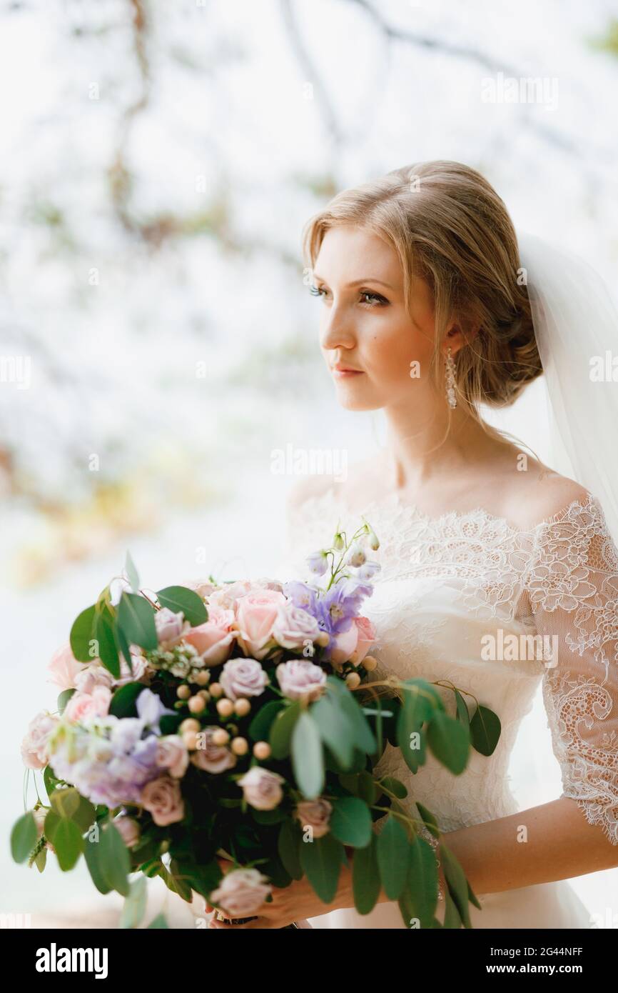 Una sposa sofisticata si erge con un bouquet di nozze tra le mani sotto i rami di un albero, primo piano Foto Stock