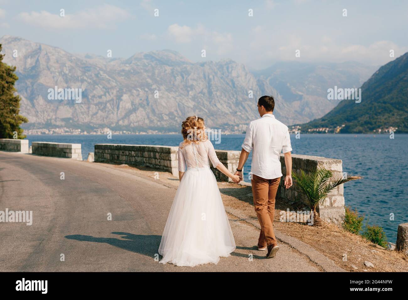 La sposa e lo sposo camminano mano in mano lungo la strada lungo la costa nella baia di Cattaro vicino Perast, vista posteriore Foto Stock