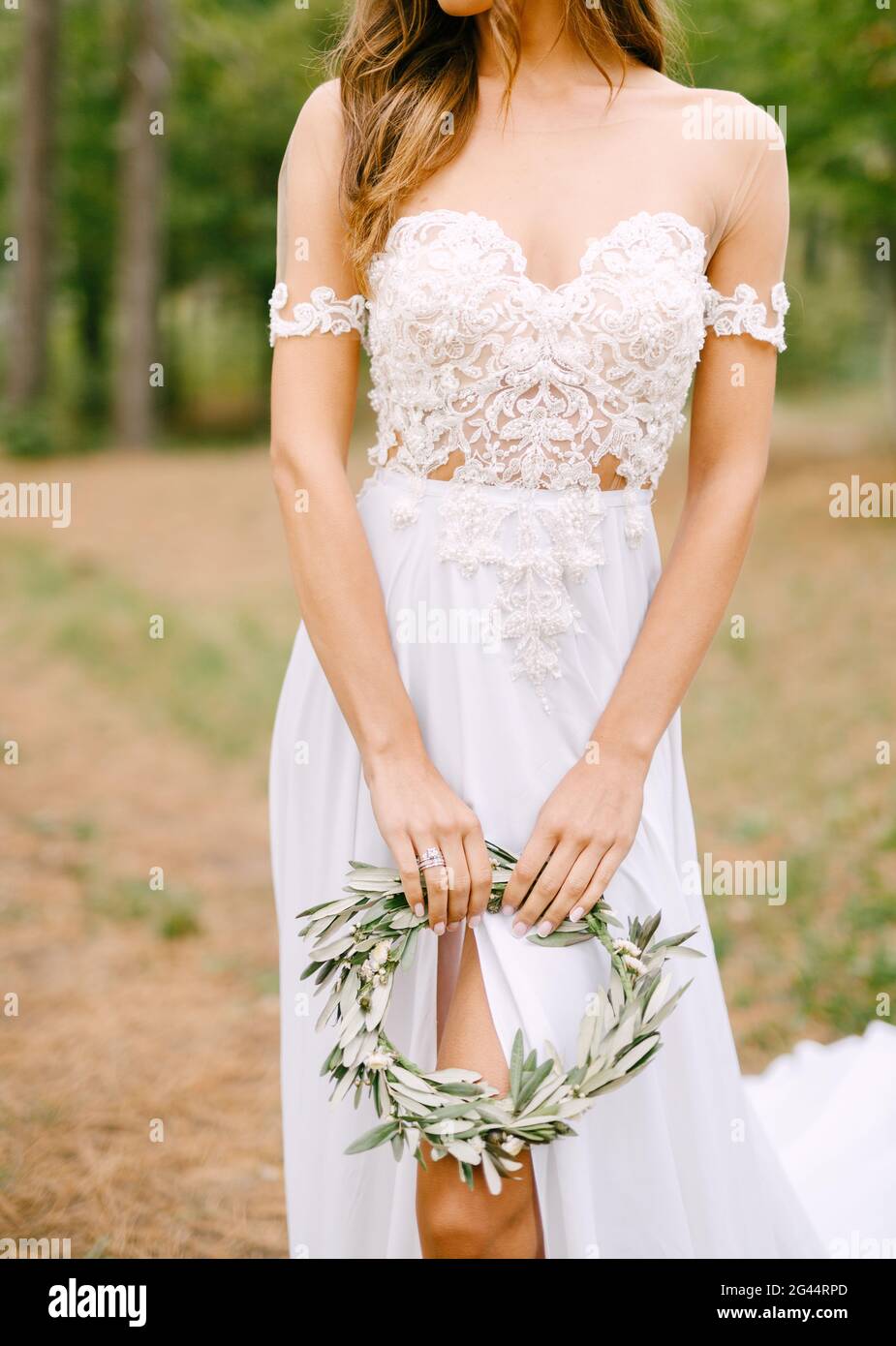 Elegante sposa in un abito ricamato in pizzo con un taglio sulla gamba che regge una corona di rami di olive Foto Stock