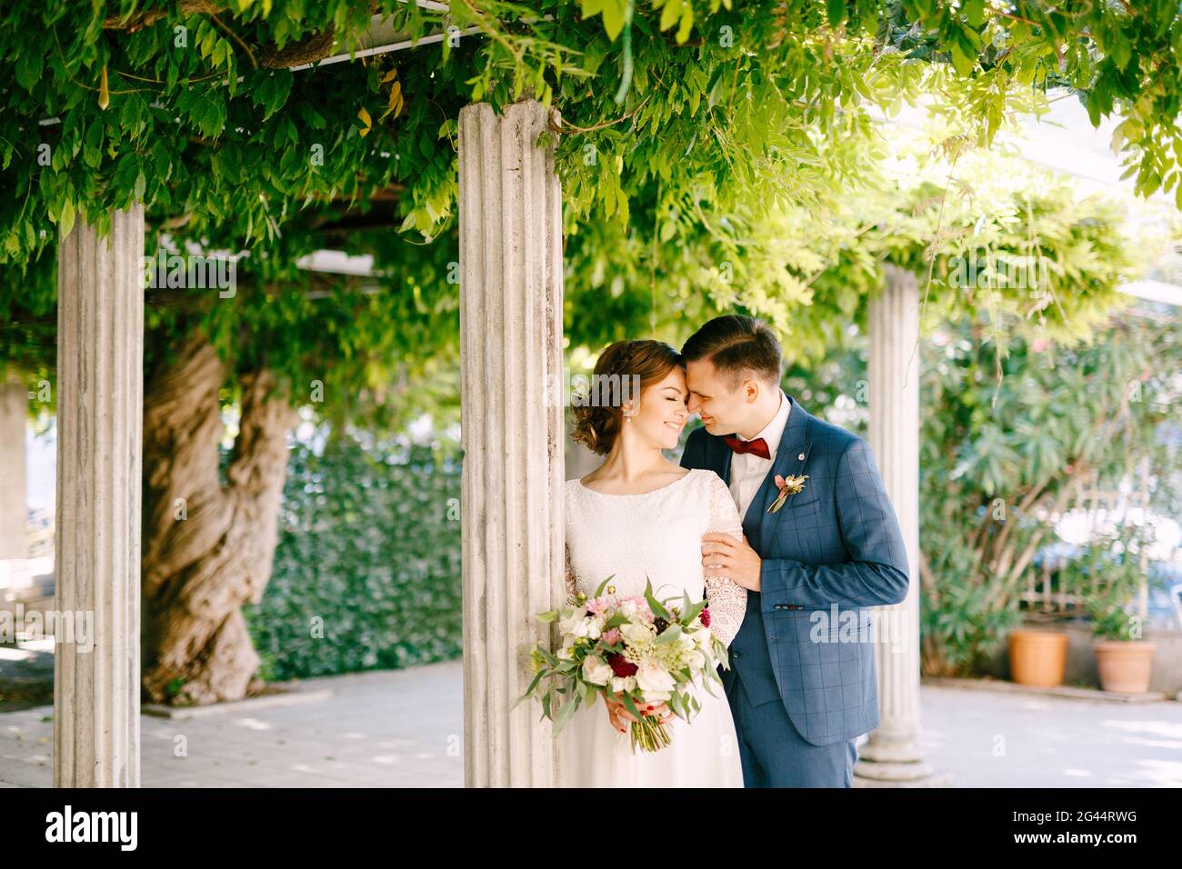 Lo sposo sorridente abbraccia le spalle della sposa sullo sfondo di colonne in un giardino verde Foto Stock