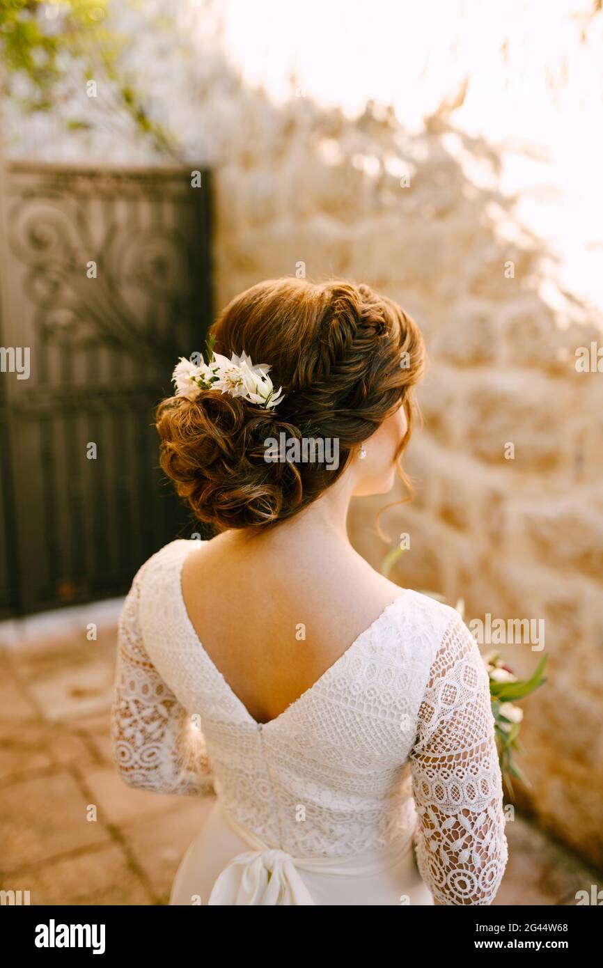 Elegante sposa si erge con un abito in pizzo bianco con un'acconciatura di lusso e fiori nei suoi capelli guarda il muro di fronte Foto Stock