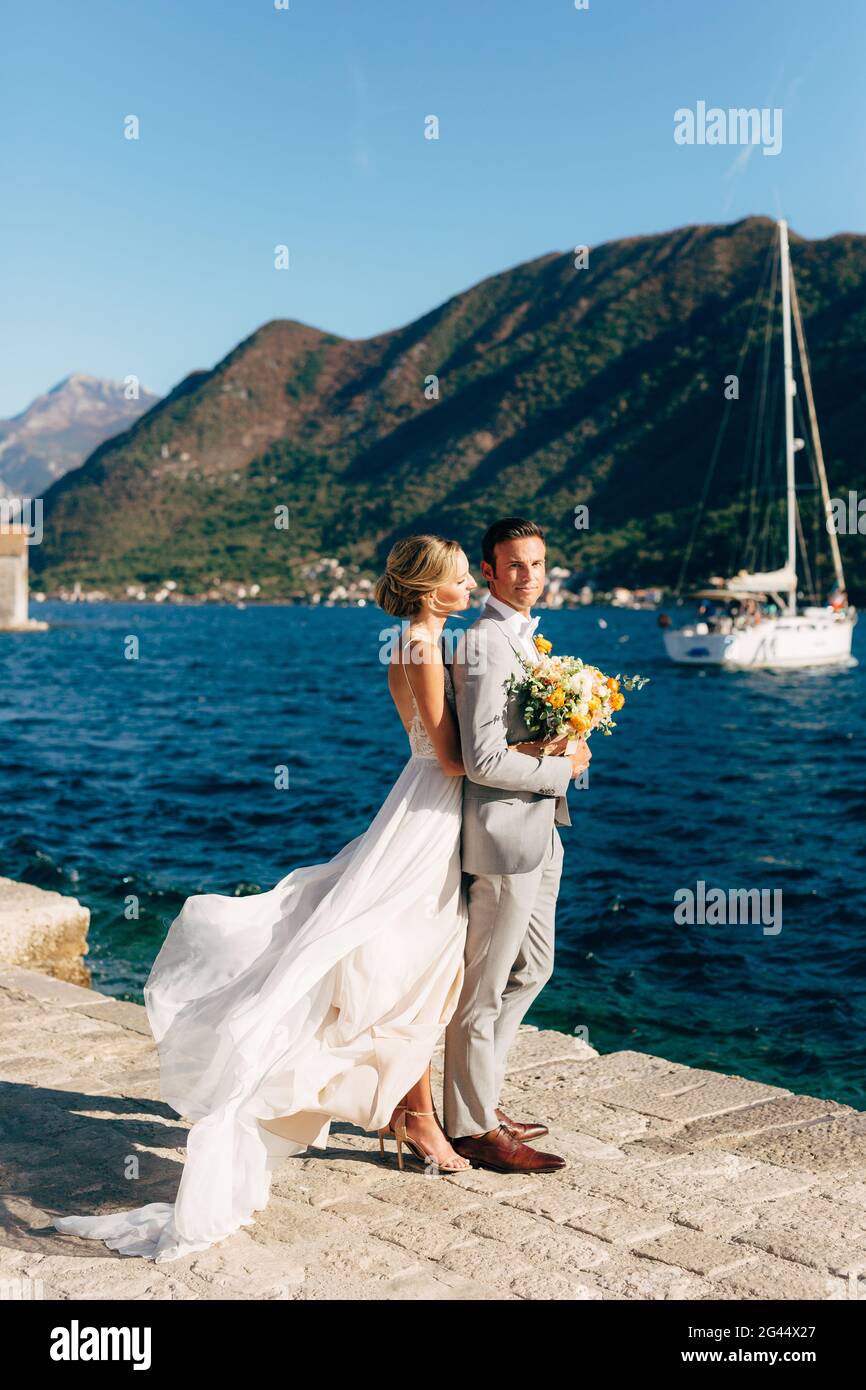 La sposa e lo sposo si abbracciano sul molo nella Baia di Cattaro, dietro di loro ci sono montagne verdi e uno yacht bianco Foto Stock