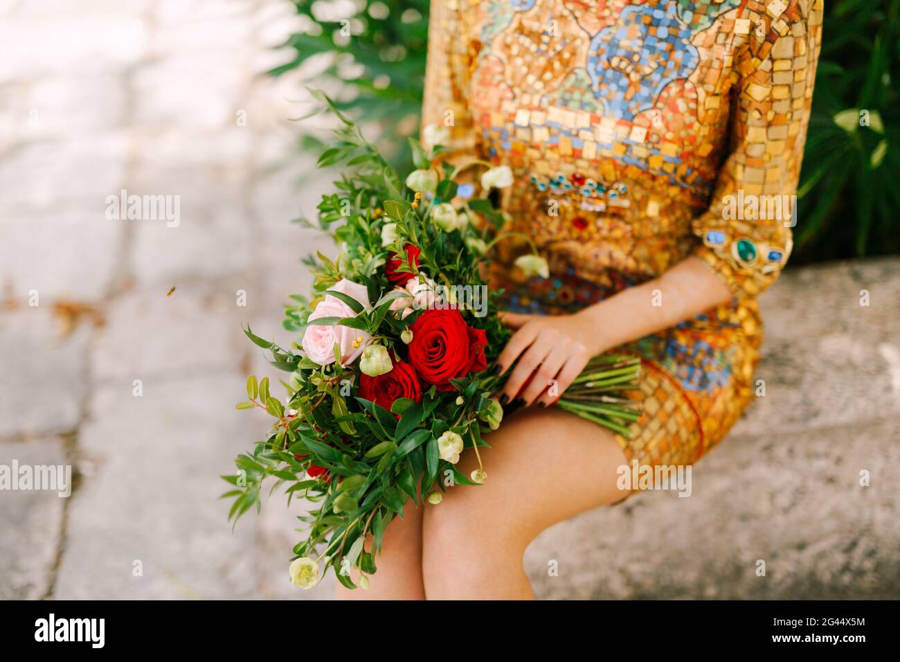 Una sposa in un insolito abito d'oro con icona si siede e tiene un bouquet nuziale di rose bianche e rosse nelle sue mani, primo piano Foto Stock