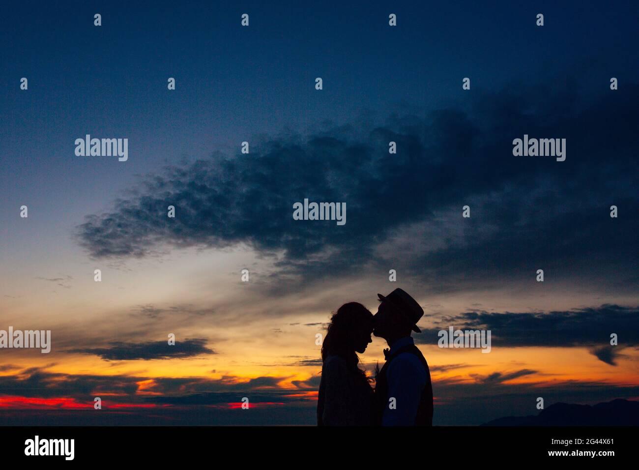 La silhouette dello sposo nel cappello bacia sposa sul fronte al tramonto Foto Stock