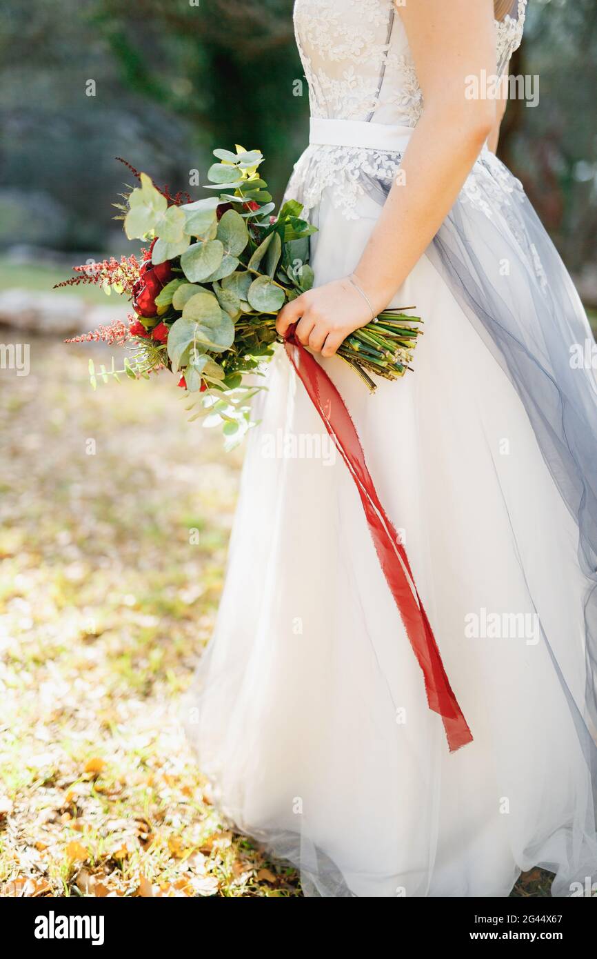 Una sposa in un abito da sposa grigio chiaro tiene un bouquet con peonie rosse, astilba, rose e eringio e nastri lunghi rossi, close-u Foto Stock