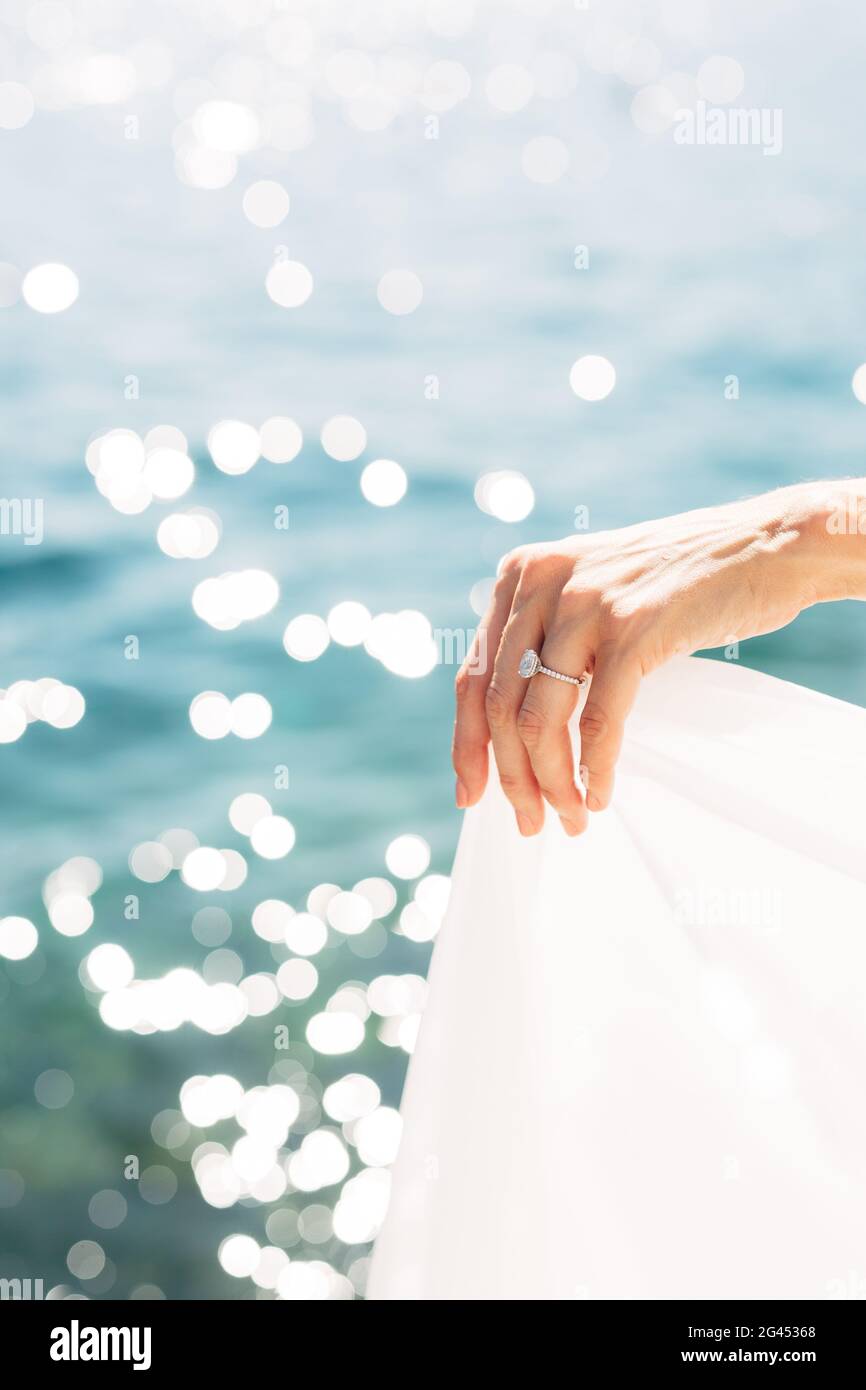 La mano della sposa con un anello di nozze sul suo dito, primo piano. La sposa tiene il bordo del vestito contro lo sfondo del se Foto Stock