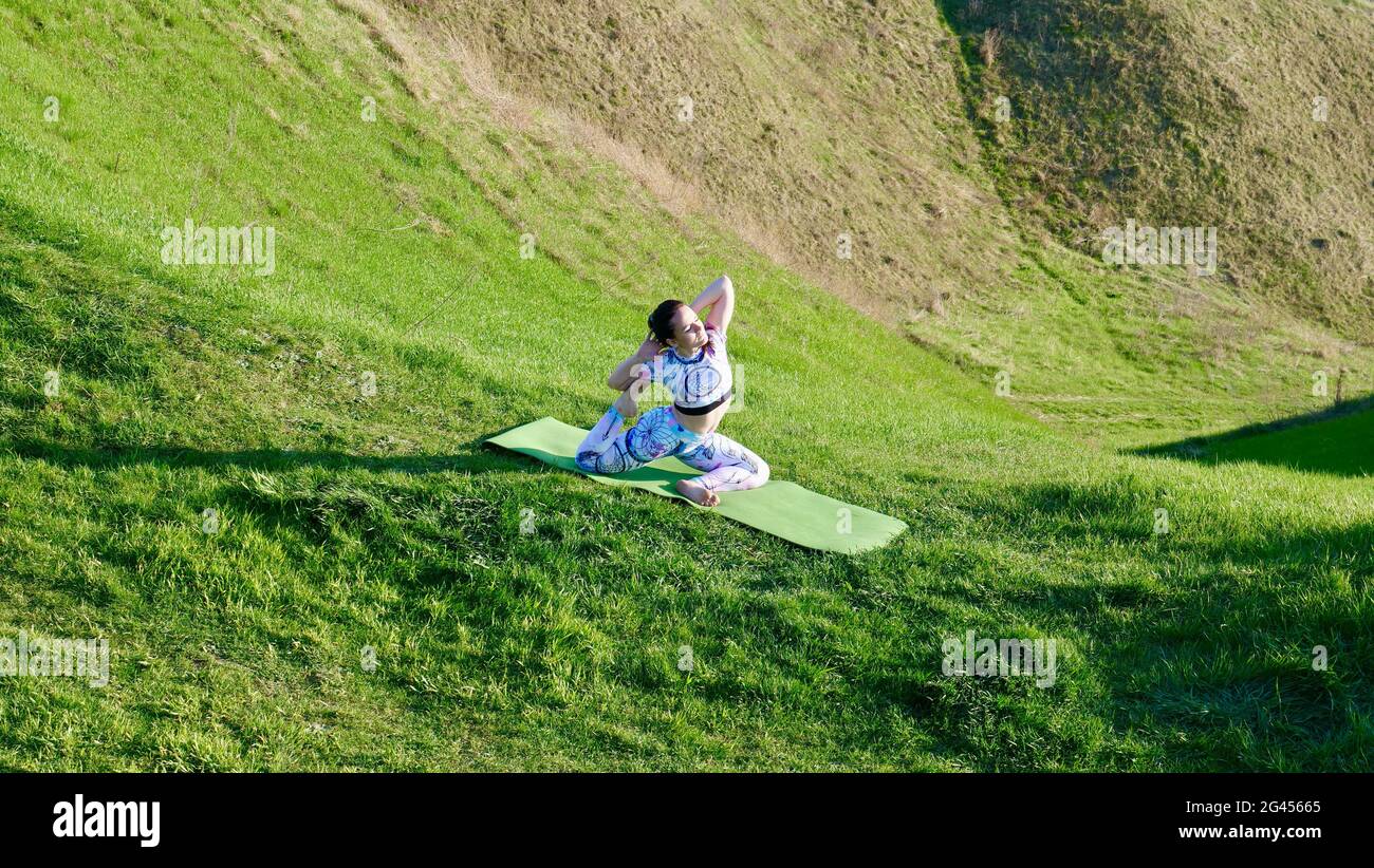 Donna yogi praticare yoga in natura, verdi colline in estate su un tappetino yoga. Una ragazza caucasica dai capelli castani in un'asana in abbigliamento sportivo, leggings e una T-shirt con capelli intrecciati in una coda di pony. Foto Stock