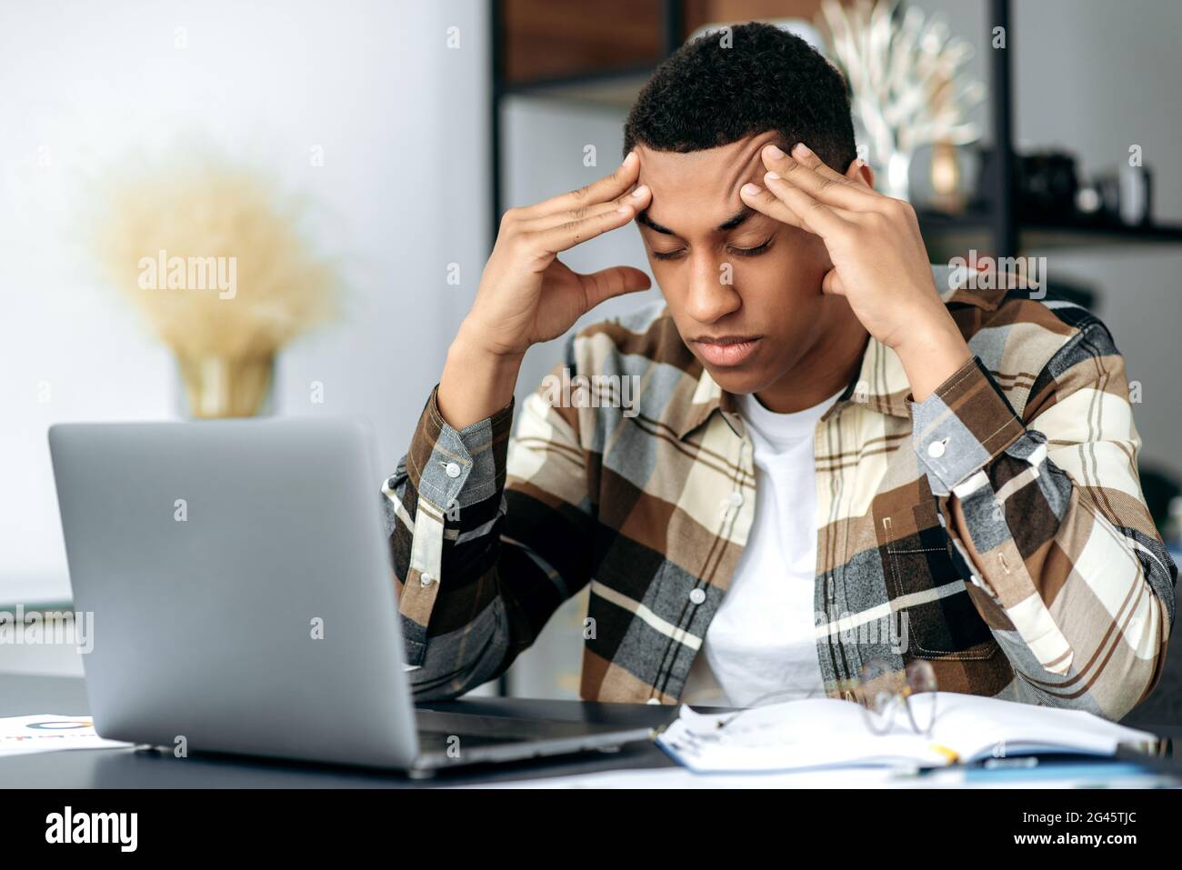 Depresso giovane latino stanco, in abiti casual eleganti, freelance, seduto alla sua scrivania, massaggiando i suoi templi con gli occhi chiusi, pensando a un progetto, hanno un mal di testa, emicrania, overwork Foto Stock