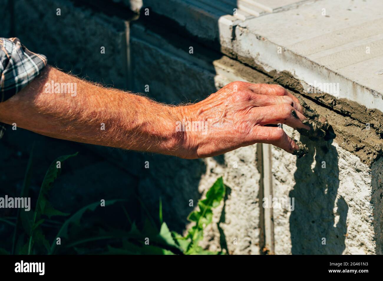 Un vecchio muratore stende le piastrelle di calcestruzzo all'esterno in una giornata estiva. Mano maschio con Malta cementizia primo piano. Foto Stock