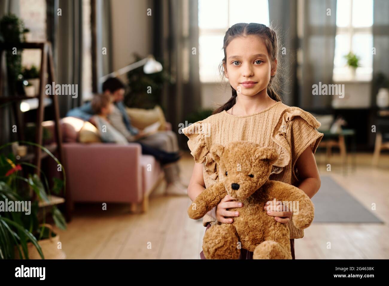 Ragazza carina con orsetto in piedi in soggiorno contro la sua famiglia Foto Stock
