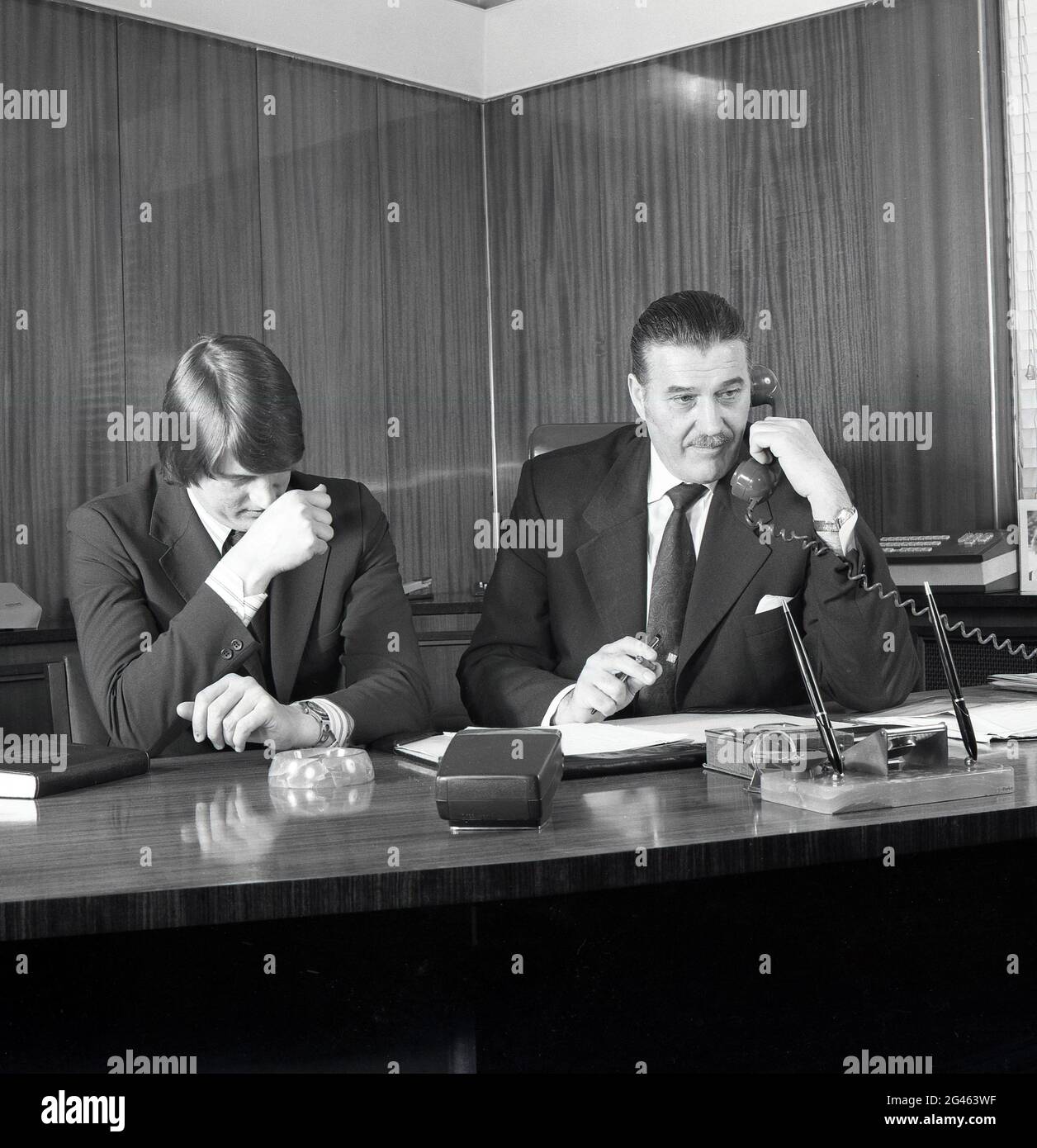 1975, storico, all'interno di un ufficio di un concessionario di automobili, un direttore della società seduto in una grande scrivania al telefono, con il suo direttore vendite accanto a lui, Croydon, Londra del Sud, Inghilterra, Regno Unito. Foto Stock