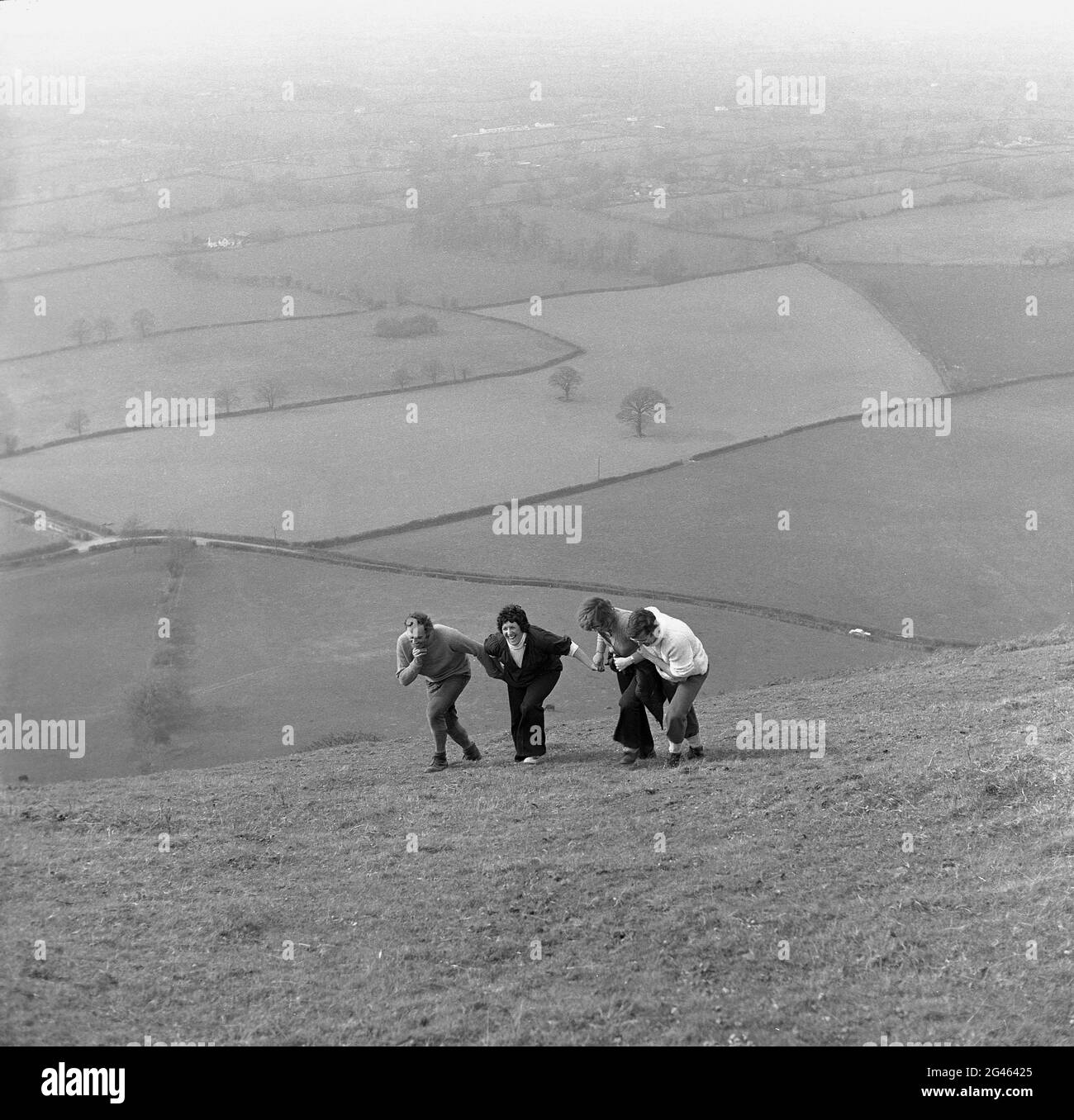 1976, storico, divertente su una collina, due coppie fuori camminando, tenere l'uno sull'altro nei loro tentativi di salire o salire su una collina erbosa ripida, Shropshire, Inghilterra, Regno Unito. Un'area montana, le colline di Shropshire è una delle regioni naturali dell'Inghilterra e un'area di eccezionale bellezza naturale (AONB). Foto Stock